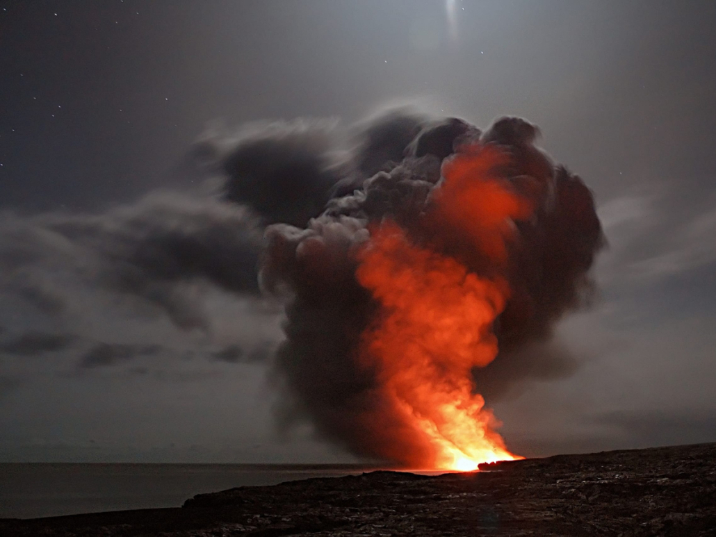 Lava erupts from a volcano on the Big Island of Hawaiʻi, sending steam and smoke into the sky near the ocean at sunset.