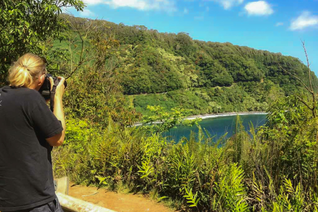 road-to-hana-visitor-photographing-cliffs-maui Tourist taking a photo of the scenic cliffs and lush greenery along the Road to Hana coastline in Maui, Hawaii.