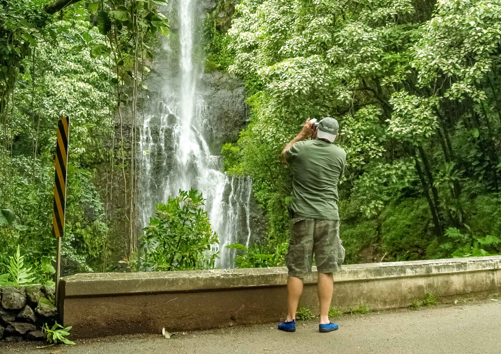 Visitor Photographing Waterfall on the Road to Hāna, Maui A visitor stops to photograph a scenic waterfall along the lush and winding Road to Hāna on Maui, surrounded by tropical greenery.